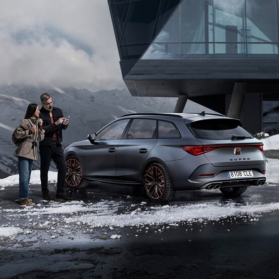 A man and woman stand beside a CUPRA Leon Sportstourer in a snowy, high-altitude setting. The man holds a smartphone, and they look at it together. The car is parked in front of a striking, modern angular building, with a vast snow-covered mountain range under a cloudy sky.