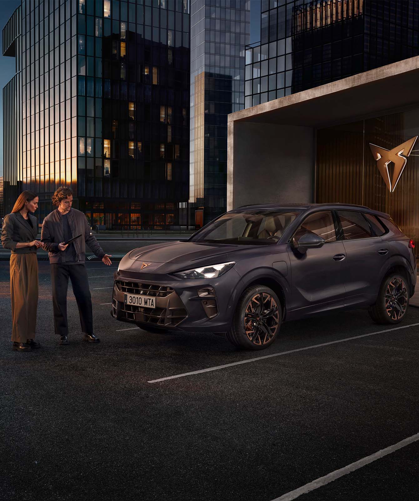 A dark void coloured CUPRA Terramar, parked in front of a modern glass building, with two people standing beside the car in an urban setting.